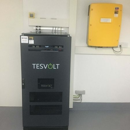 Schwarzer TESVOLT-Batteriespeicher neben einem gelben wandmontierten Wechselrichter in einem Reinraum.