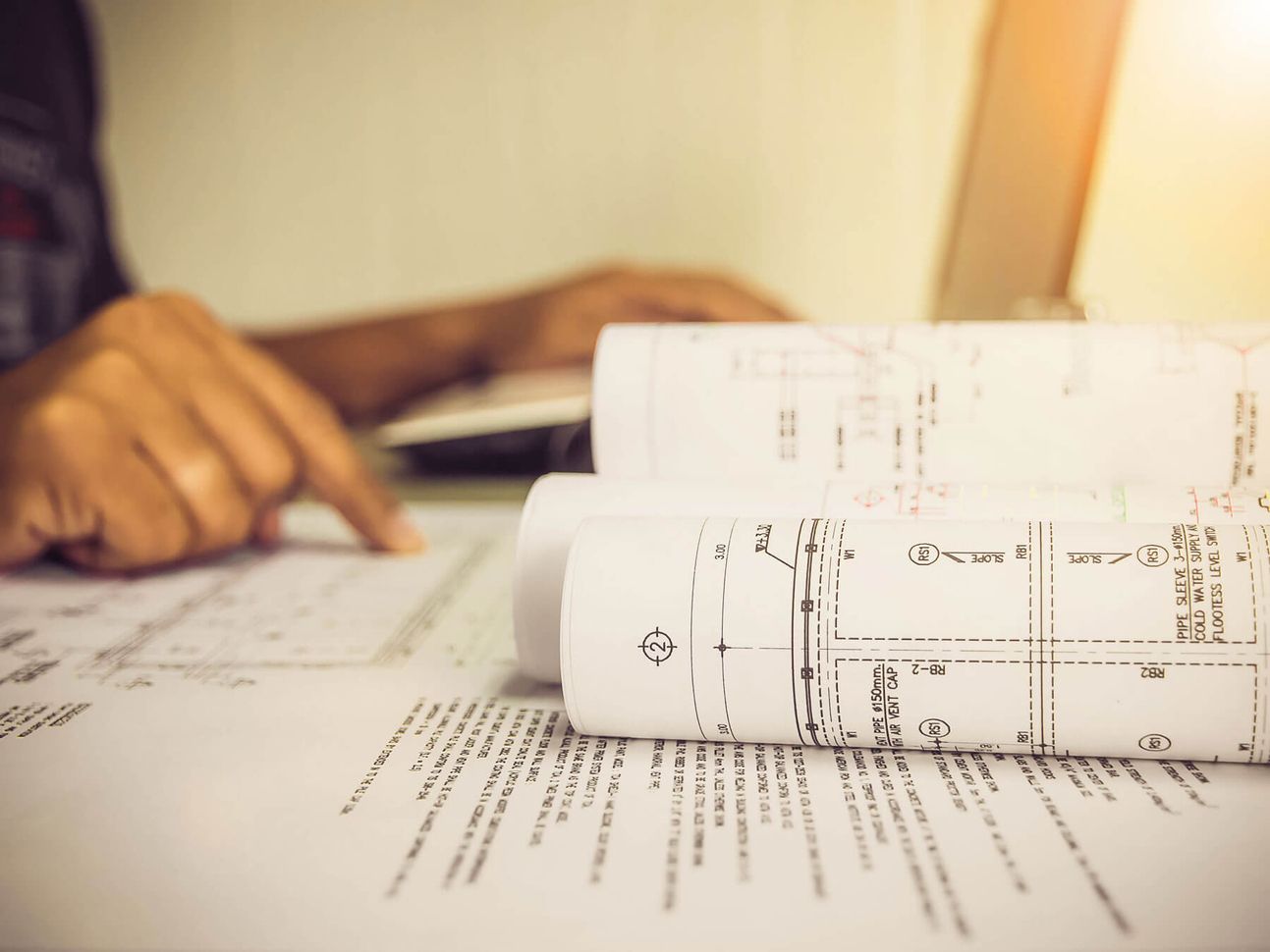 Person, die an einem Schreibtisch mit warmer Beleuchtung Architekturpläne prüft.